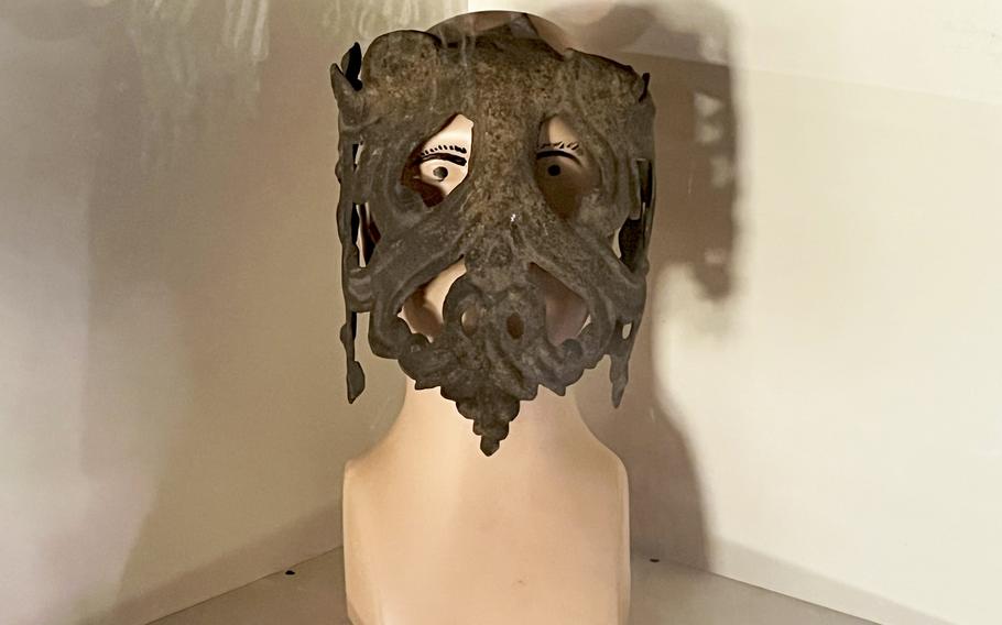 Close-up of an ornate mask displaying intricate craftsmanship with visible eye holes, mounted on a light-colored display stand.