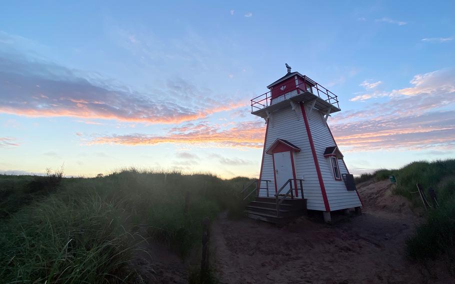 A lighthouse stands among grassy dunes with the sun setting over the horizon in the background.