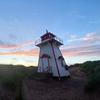A lighthouse stands among grassy dunes with the sun setting over the horizon in the background.