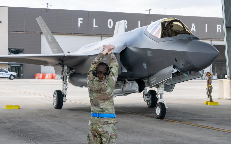 A service member signals to a pilot seated in the cockpit of a fighter jet on a runway.