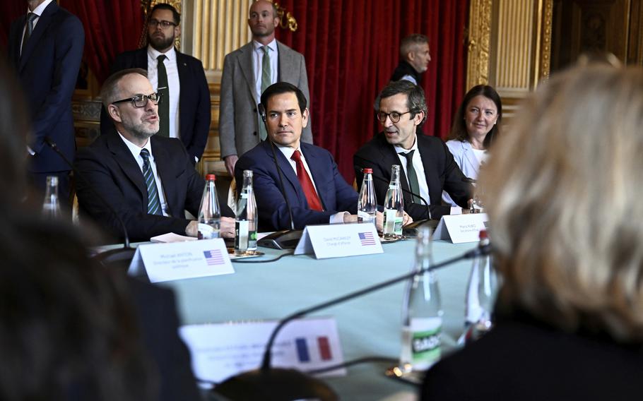 Marco Rubio in the center of a photo, sitting at a table with other people in suits on either side of him.