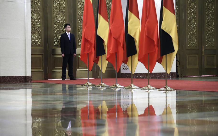 German and Chinese flags on the top half of an image with a security officer standing next to the flags and the flags’ reflection on a polished floor taking up the bottom half of the image.