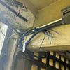 Interior view of a room showing an exposed ceiling with peeling paint, electrical conduit and multiple blue wires running along the walls with frayed ends next to a small white electrical box.