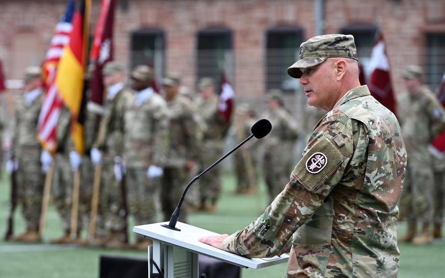 The side profile of a man in an Army uniform speaking into a microphone as other soldiers stand in formation in the background.