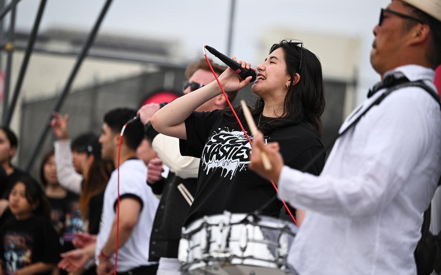A woman in a black T-shirt sings into a microphone on stage with a drummer to the side and other musicians behind her.