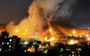 Smoke and fires rise from a complex near a hill at night.