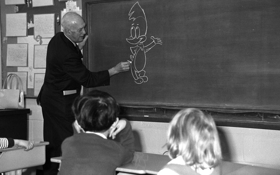 A black and white photo of a man in a suit drawing cartoon birds on a blackboard as children watch from their desks.