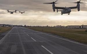 Three MV-22 Osprey tiltrotor aircraft fly low over a runway during sunset, with buildings and grassy fields on either side of the airstrip under a cloudy sky.