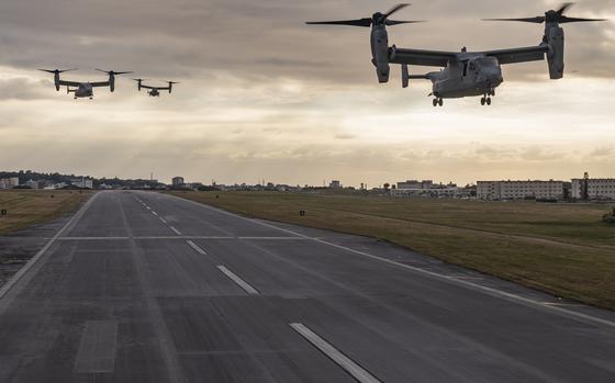 Three MV-22 Osprey tiltrotor aircraft fly low over a runway during sunset, with buildings and grassy fields on either side of the airstrip under a cloudy sky.