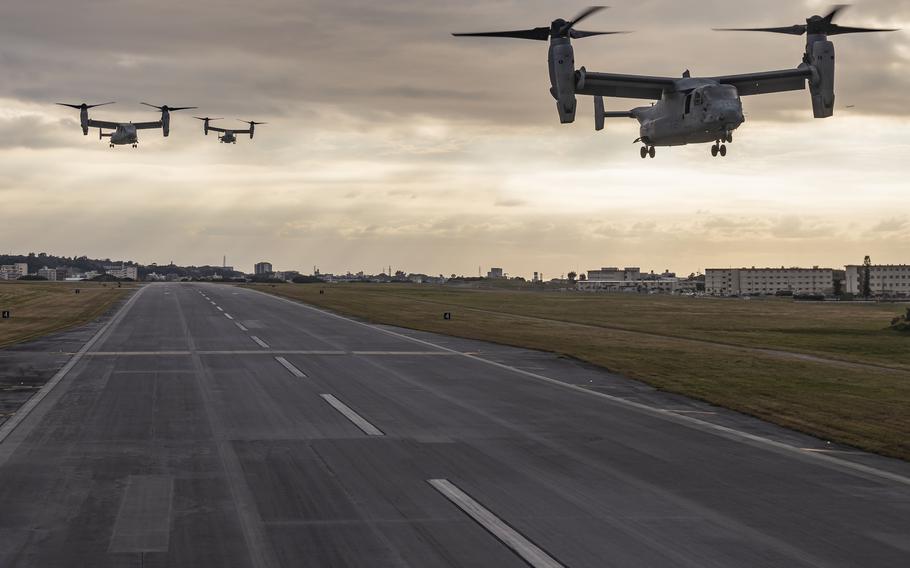 Three MV-22 Osprey tiltrotor aircraft fly low over a runway during sunset, with buildings and grassy fields on either side of the airstrip under a cloudy sky.