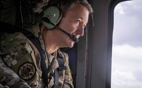 A man in a camouflage uniform looks out an aircraft window. He wears a communication headset with microphone and has military patches visible on his uniform.