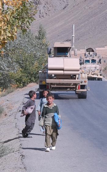 Three small children walking on a road with two military vehicles driving behind them.