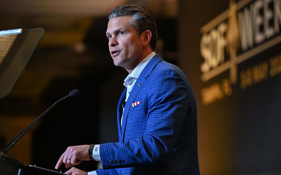 Pete Hegseth, wearing a blue blazer and seen from the side, stands at a podium and speaks into a microphone.