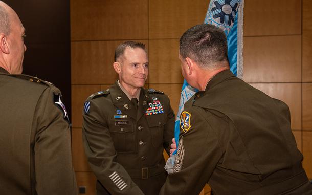 Three men in formal Army uniforms, two of them exchanging a flag.