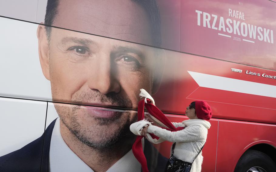 A woman stands next to a red campaign poster with the face of a Polish presidential candidate.