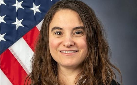 A woman with brown hair, seen from the neck up, poses for a portrait in front of a U.S. flag.