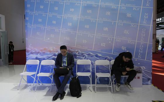 Two men sit on chairs before a poster of a periodic table, one in a suit working on a laptop and the other in casual clothes looking at his phone.