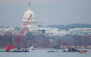 A large red crane, seen from a distance, lifts wreckage of a crashed aircraft from water with the U.S. Capitol building and city skyline in the background.