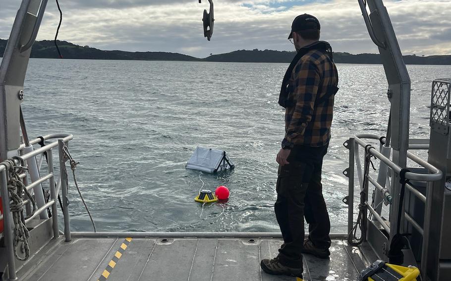 A person in a plaid flannel shirt and baseball cap stands on the deck of a research vessel, looking out over calm water under a cloudy sky. Scientific equipment including a white pyramid shaped floating reflector and a red buoy float in the water nearby, connected by cables to the boat. 