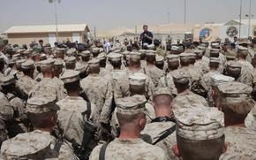 Rows of service members stand and listen to a man speaking into a microphone in the distance.