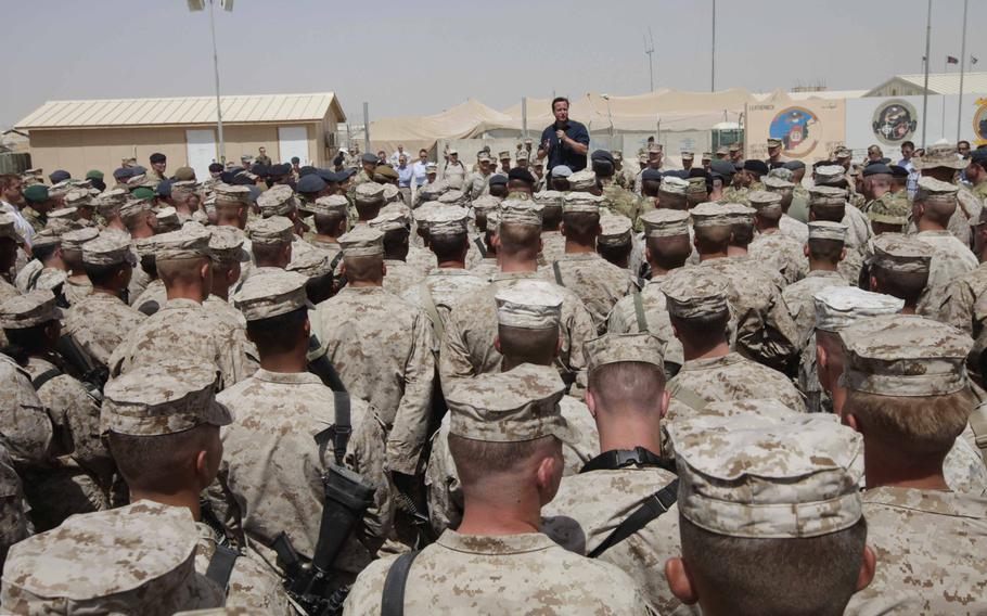 Rows of service members stand and listen to a man speaking into a microphone in the distance.
