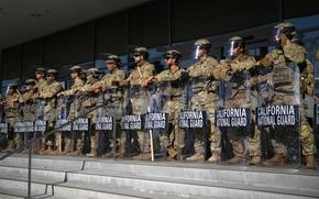 Military troops in camouflage uniforms and helmets stand on stone steps in front of the entrance to a government buildings while leaning on riot shields that read “California National Guard.”