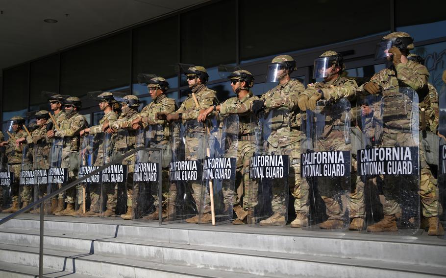 Military troops in camouflage uniforms and helmets stand on stone steps in front of the entrance to a government buildings while leaning on riot shields that read “California National Guard.”