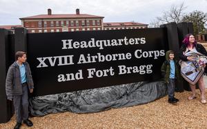 A woman and two boys stand on either side of a brown welcome sign reading “Headquarters XVIII Airborne Corps and Fort Bragg.”