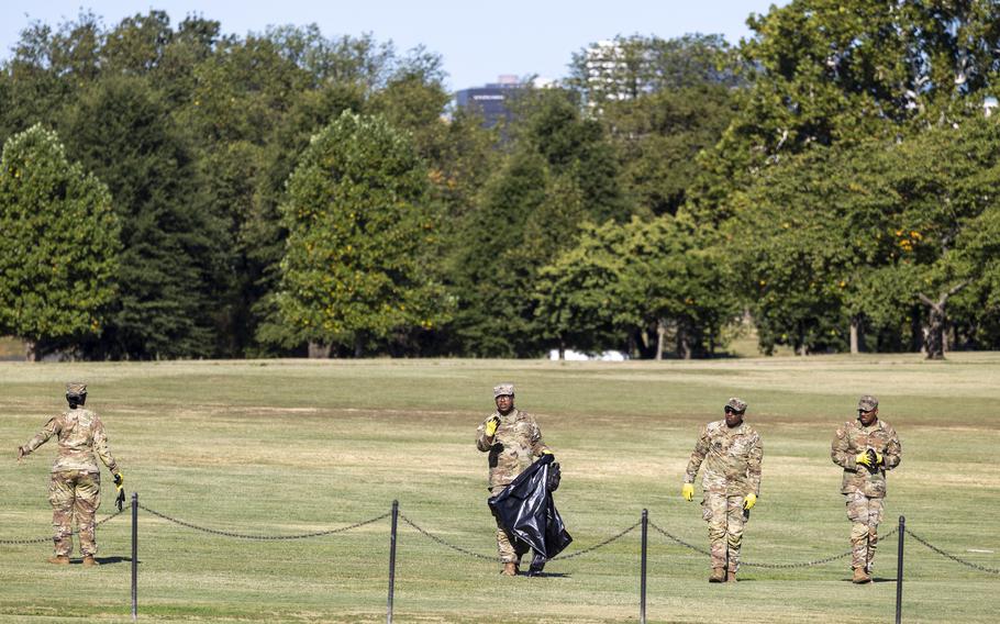 Four soldiers walking on a large lawn, one holding a black trash bag.