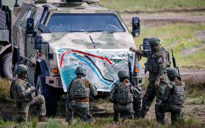 Military personnel in camouflage uniforms gathered around an armored vehicle, examining a large topographical map spread across the front of the vehicle. The soldiers are equipped with helmets and tactical gear, positioned in a grassy field.