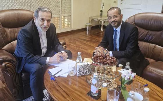 Two men in dark suits sit in brown leather chairs around a coffee table with documents, bottles of water, glasses and a centerpiece with food on it.