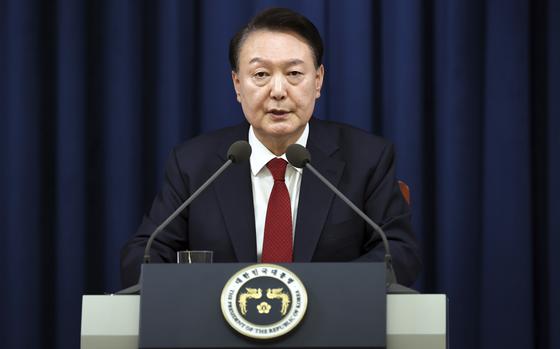 A Korean man in a dark suit sits at a lectern and speaks into two microphones in front of a blue curtain in the background.