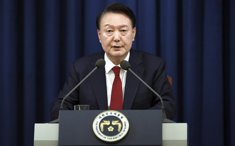 A Korean man in a dark suit sits at a lectern and speaks into two microphones in front of a blue curtain in the background.