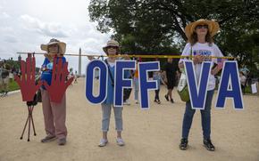 Three women stand on a dirt path holding up a sign reading “Hands off VA,” with the Washington Monument in the background.