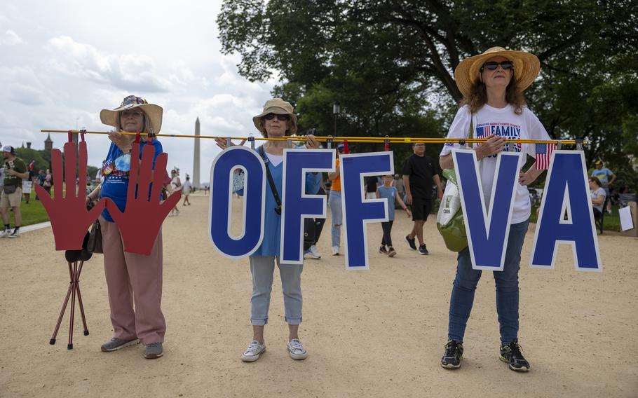 Three women stand on a dirt path holding up a sign reading “Hands off VA,” with the Washington Monument in the background.