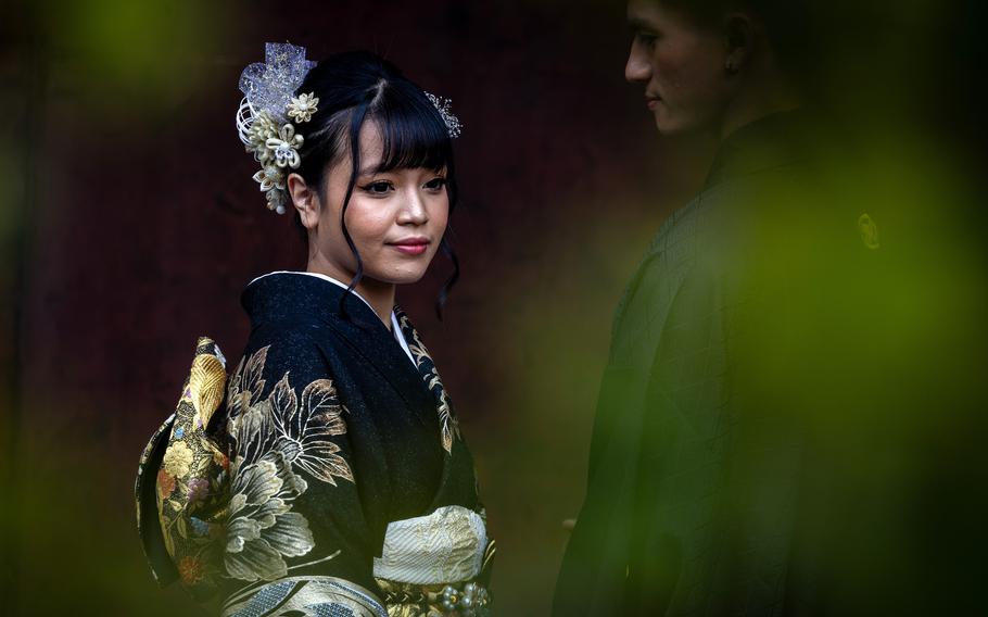 A Japanese woman wearing a black kimono with flower patterns and a flower in her hair stands next to a Japanese man in a traditional robe obscured by greenish haze.