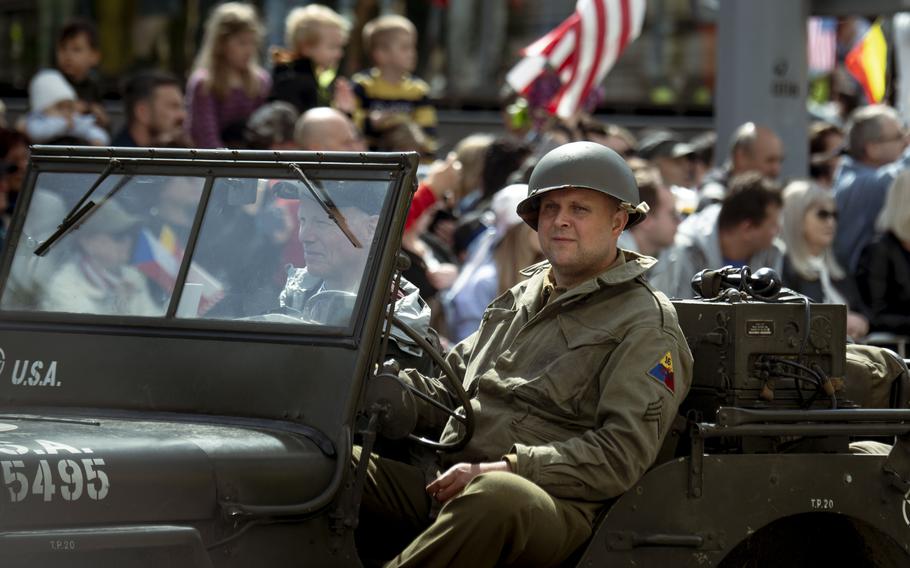 A man in uniform in a jeep rolls past crowds and an American flag.