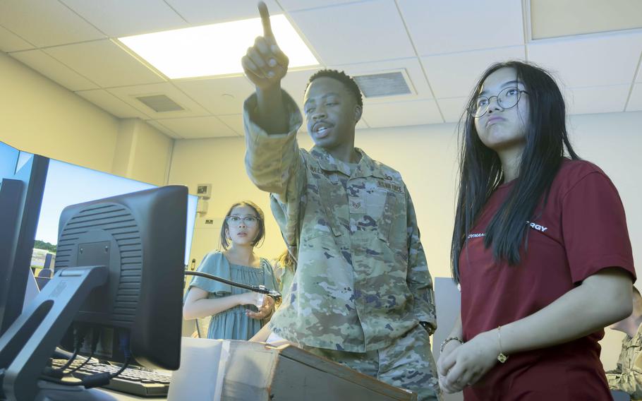 An airman is pointing at a screen while explaining something to a group of students inside a well-lit classroom or office. One student in a red shirt stands beside him, attentively listening, while another student in a green dress looks on from the background. Computer monitors and other equipment are visible on the desk in front of them.