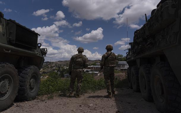 Two soldiers in camouflage combat gear and helmets, seen from behind, stand between armored vehicles as they look out onto a desert landscape under a cloudy, blue sky.
