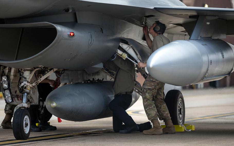 Military personnel in camouflage uniforms perform maintenance on a fighter jet, with one service member working on ordnance mounted under the aircraft’s wing while another tends to the landing gear area on an airfield tarmac.