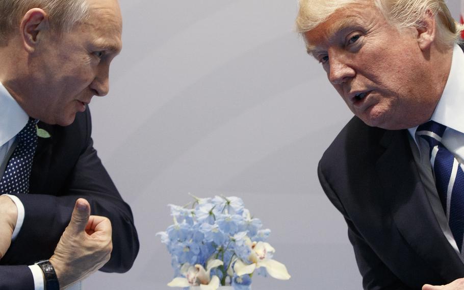 Vladimir Putin and Donald Trump lean in to speak to each other over a table with a vase of flowers on it.