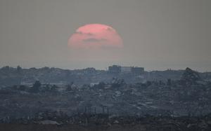 The sun, cast in a pinkish hue and obscured by clouds, sets over a skyline of destroyed buildings.