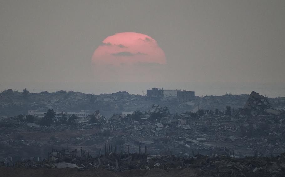 The sun, cast in a pinkish hue and obscured by clouds, sets over a skyline of destroyed buildings.