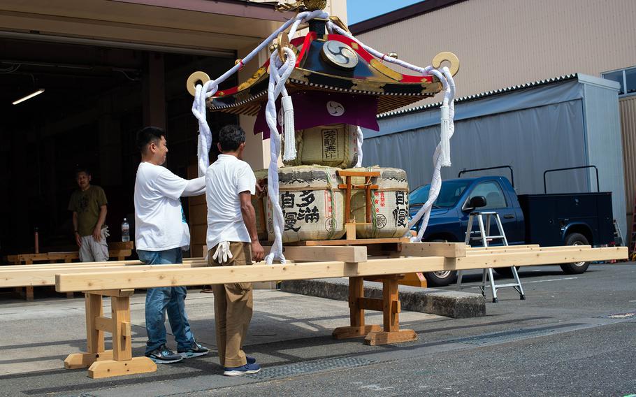 Two men in white shirts examine a portable Japanese shrine (mikoshi) that sits on wooden supports. The shrine is decorated with white ropes, traditional ornaments and sake barrels.