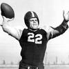 A black and white photo of a football player with both hands raised, one gripping a football.