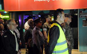 A group of men, including military personnel in camouflage uniforms, police officers and civilians, stand in formation on a sidewalk at night in front of a brightly lit bar with a red sign. One man in the foreground wears a reflective safety vest. The atmosphere suggests a coordinated event or patrol, possibly related to community safety or a formal inspection. 