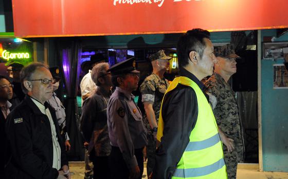 A group of men, including military personnel in camouflage uniforms, police officers and civilians, stand in formation on a sidewalk at night in front of a brightly lit bar with a red sign. One man in the foreground wears a reflective safety vest. The atmosphere suggests a coordinated event or patrol, possibly related to community safety or a formal inspection. 