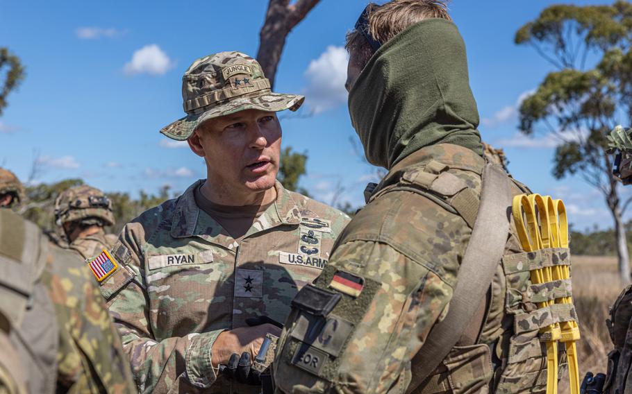 A general interacts with a soldier, who is wearing an army green scarf over his face and carrying yellow cables on his back, during an exercise. 
