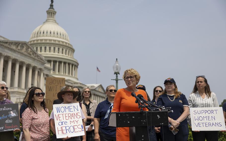 A group of women stand in front of a podium and hold signs with the U.S. Capitol in the background.
