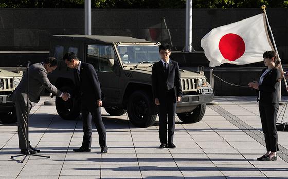 Sergiy Korsunsky, center, Ambassador of Ukraine to Japan, left, and Toshiro Ino, second left, state minister of Defense of Japan, shake hands during the handover ceremony of defense equipment for Ukraine at the Ministry of Defense Wednesday, May 24, 2023, in Tokyo. Japan Ground Self-Defense Force's the 1/2-ton trucks, seen in background, are part of handover defense equipment. (AP Photo/Eugene Hoshiko)